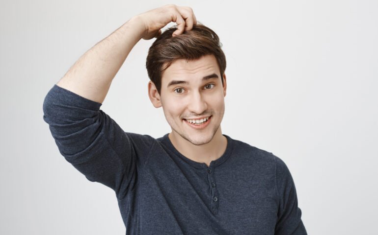 Portrait of funny handsome caucasian man in casual clothes acting like monkey, smiling and scratching head, standing against gray background. Boy attend acting classes in local theatre